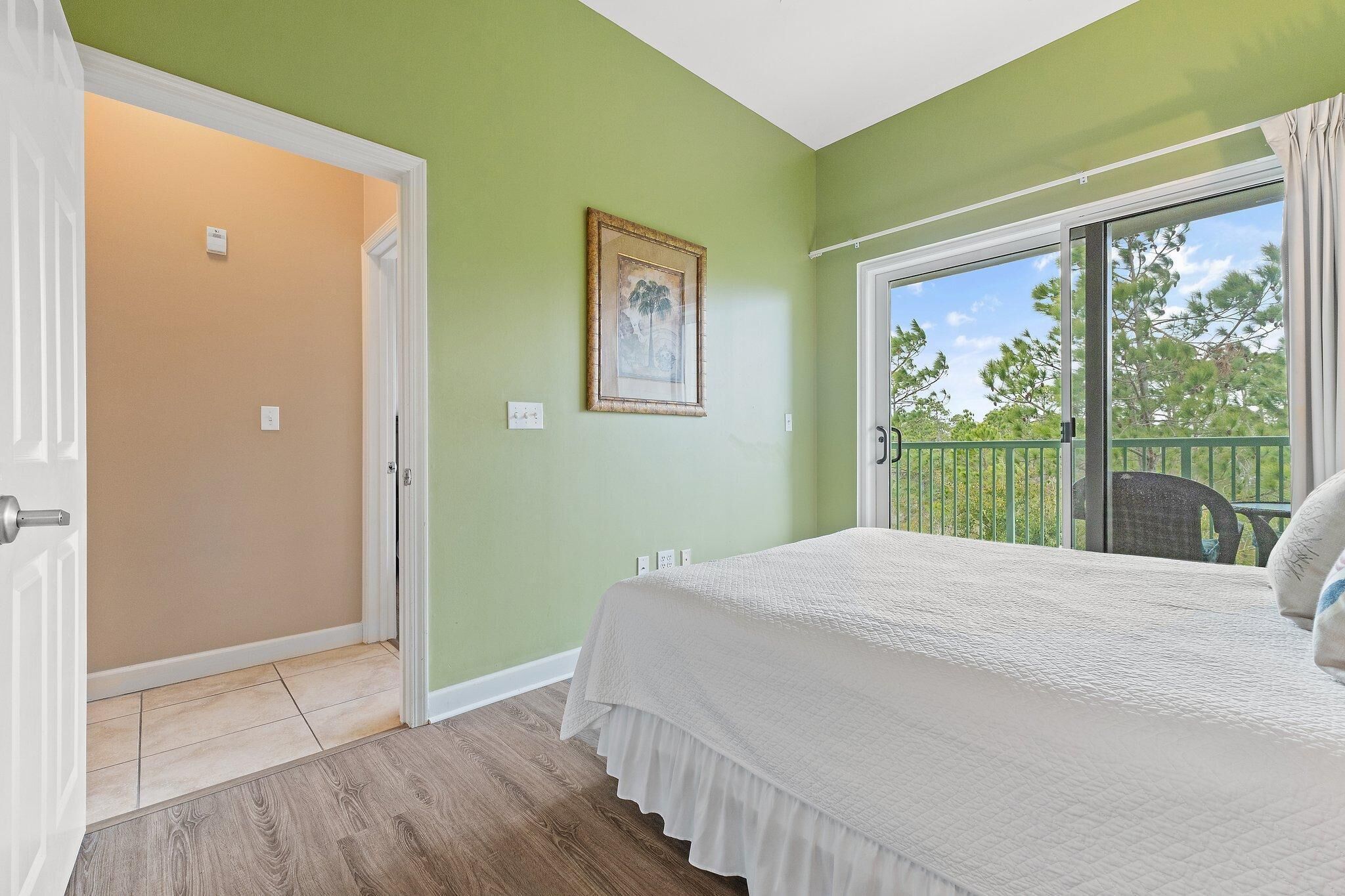 Une chambre avec un lit, une porte donnant sur une autre pièce, une fenêtre avec vue sur les arbres et un tableau encadré au mur.