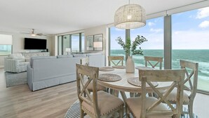 A modern living room with a large sofa, a dining table with chairs, a ceiling fan, and a large window overlooking the ocean.