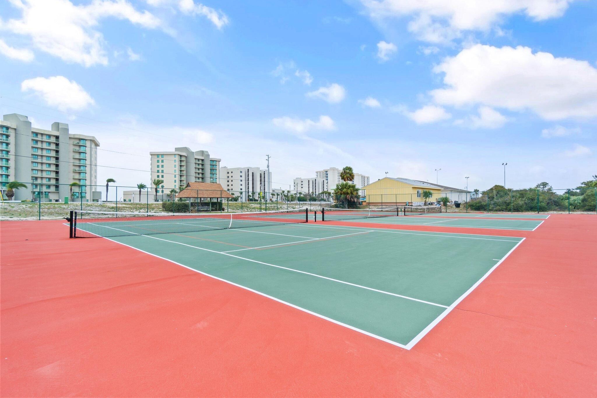 A tennis court with multiple courts, surrounded by buildings and a clear sky.