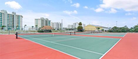 A tennis court with multiple courts, surrounded by buildings and a clear sky.