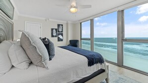 A bedroom with a large bed, a ceiling fan, and a view of the ocean through large windows.