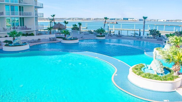 A large, curved swimming pool with multiple sections, surrounded by palm trees and lounge chairs, with a view of the sea and a building in the background. - Caribe C104 (Orange Beach)