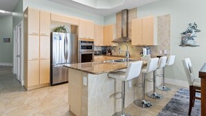 A modern kitchen with light wood cabinets, stainless steel appliances, and a central island with bar stools. - Caribe C104 (Orange Beach)