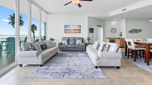 A spacious living room with two sofas, a coffee table, and a ceiling fan. A large window offers a view of palm trees and the ocean. - Caribe C104 (Orange Beach)
