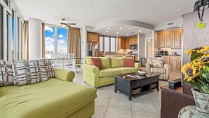A spacious living room with a green sofa, a coffee table, and a dining area with a table and chairs. The kitchen is visible in the background with wooden cabinets and a microwave.