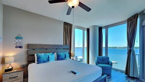 A bedroom with a large bed, a ceiling fan, a nightstand, a chair, and a view of the water.