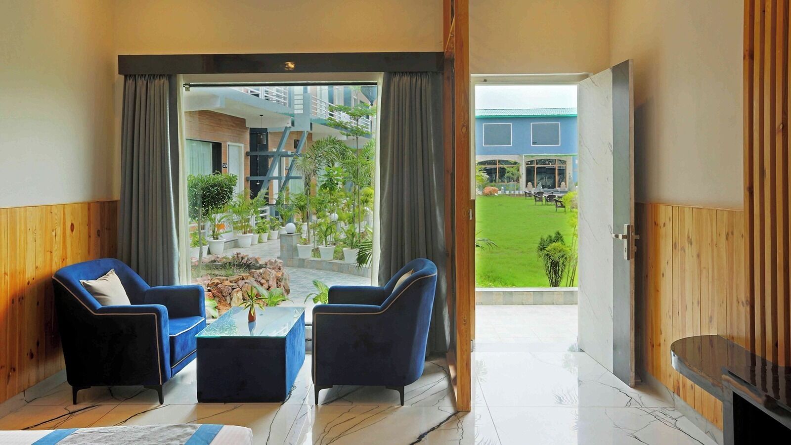 A modern room with a large window, two blue armchairs, a glass coffee table, and a view of a green garden and building.