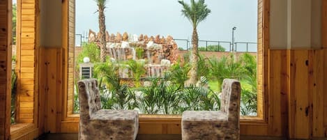A room with two plush armchairs facing a large window overlooking a landscaped garden with a waterfall and palm trees.