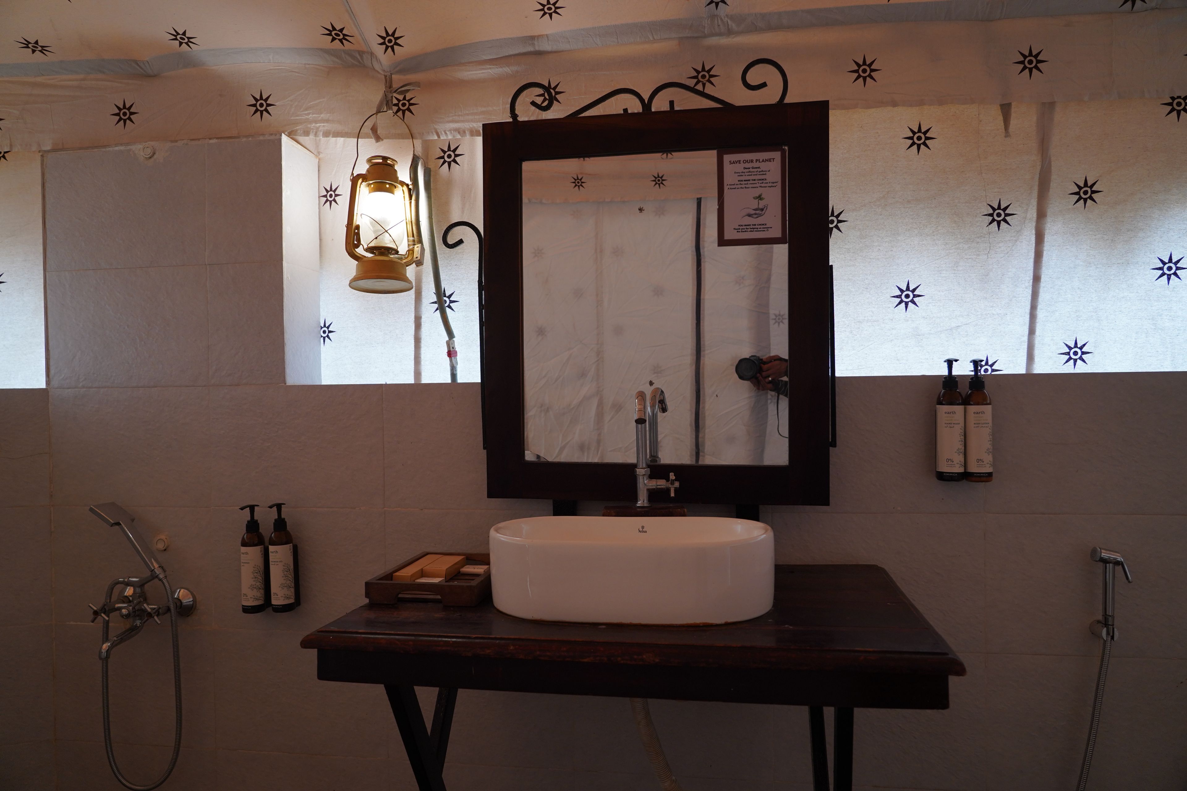 Een badkamer met een witte wastafel, een spiegel en een douche. Twee flessen Earth handzeep hangen aan de muur.
