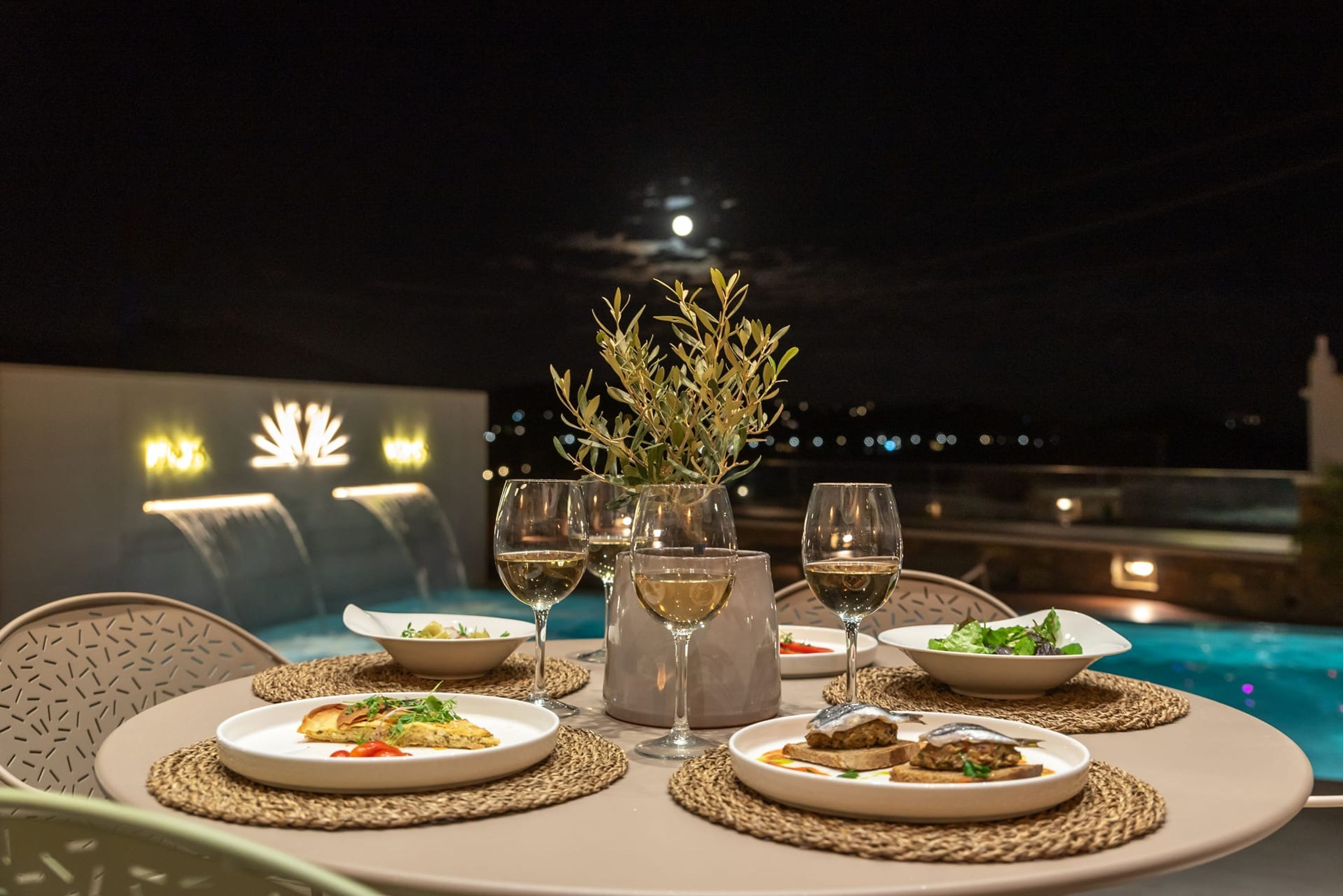 A dining table with plates of food and wine glasses, set against a night sky with a visible moon.