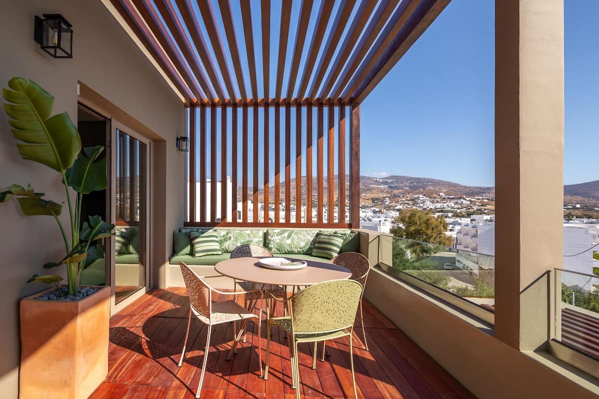 Un balcón con una pérgola de madera, una zona de estar con cojines verdes y una mesa con sillas.