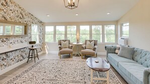 A spacious living room with a light blue sofa, two armchairs, a coffee table, and a patterned rug. A chandelier hangs from the ceiling, and there are large windows providing natural light. - ‘The Beachhouse’ Retreat w/ Prescot Bay Views! (Stockton Springs)