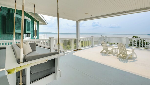 A covered outdoor seating area with a view of the water, white chairs, and a sofa with cushions.
