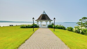 A gazebo with a tiled pathway leading to it, surrounded by green grass and flowers, overlooking a body of water.