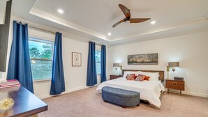 Een slaapkamer met een groot bed, een plafondventilator, een raam met gordijnen, een nachtkastje en een bijzettafeltje.