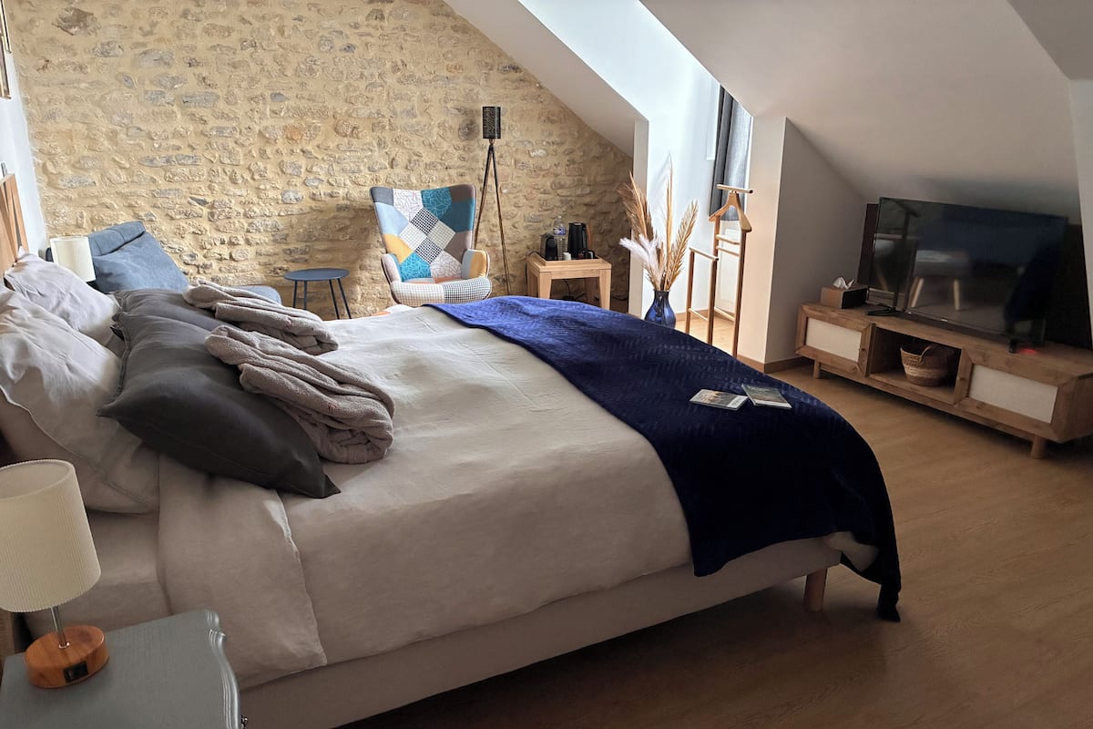A bedroom with a stone wall, a bed with multiple pillows and blankets, a small side table, a floor lamp, a wooden TV stand, and a television.