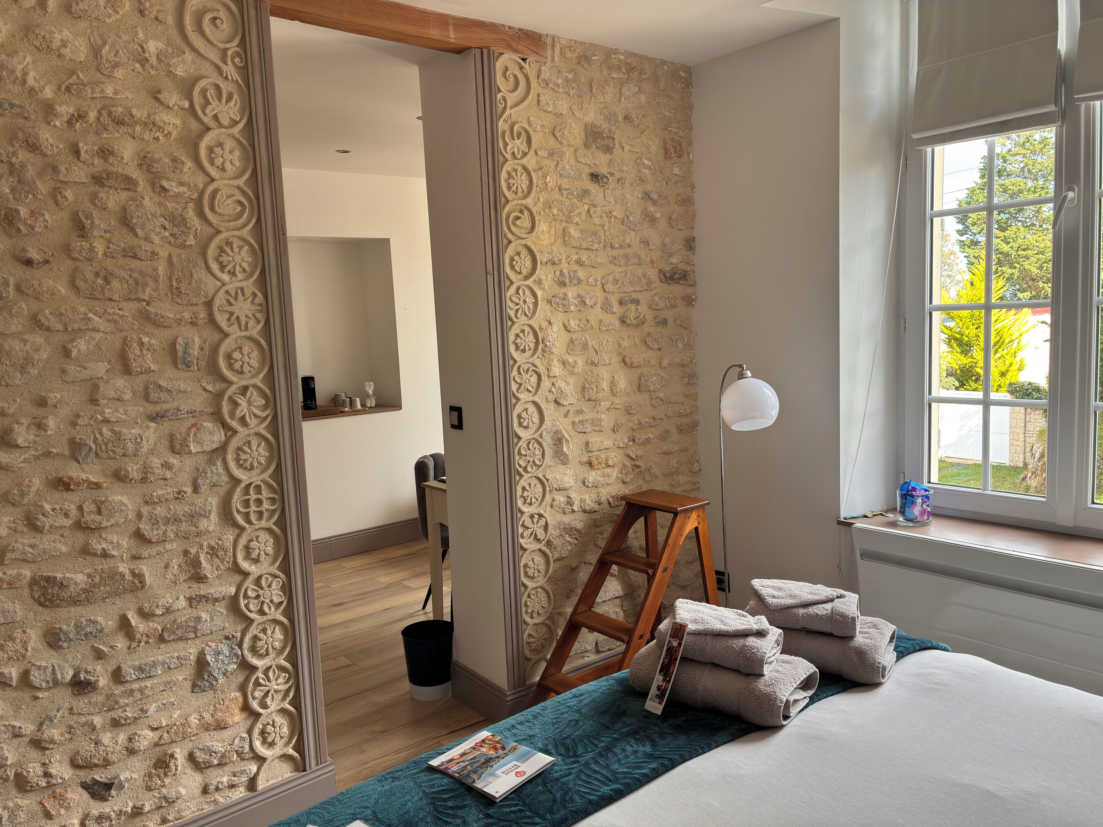 A bedroom with stone walls, a wooden ladder, a bedside table with towels, a floor lamp, and a window with a view of greenery.