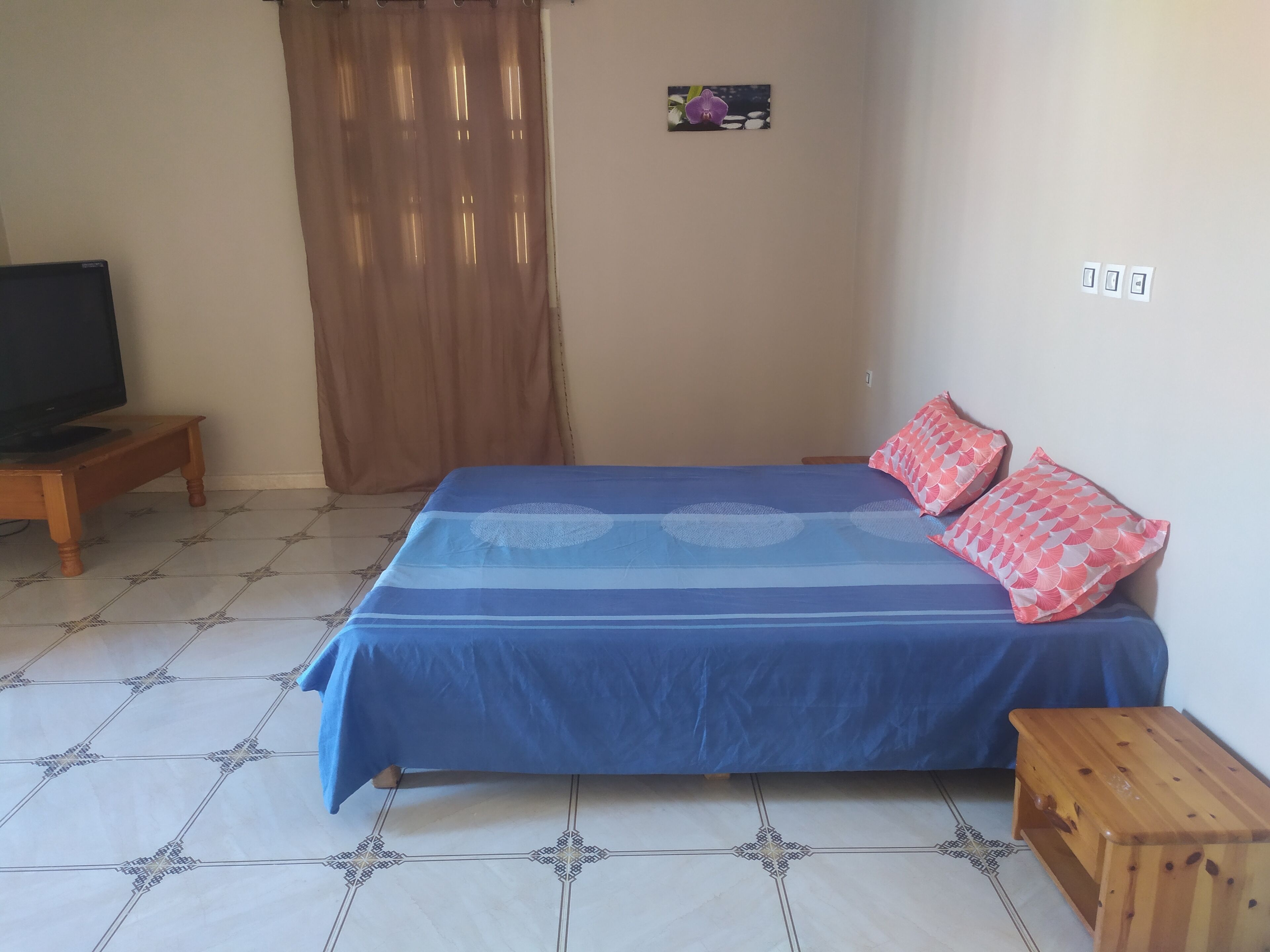 A bedroom with a bed, a television on a stand, a wooden nightstand, and a small wooden table.