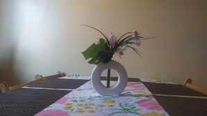 A dining table with a floral tablecloth, a white vase with a plant, and a wall sign above.