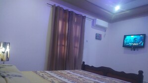 A hotel room with a bed, curtains, an air conditioning unit, a television, and a wall-mounted picture.