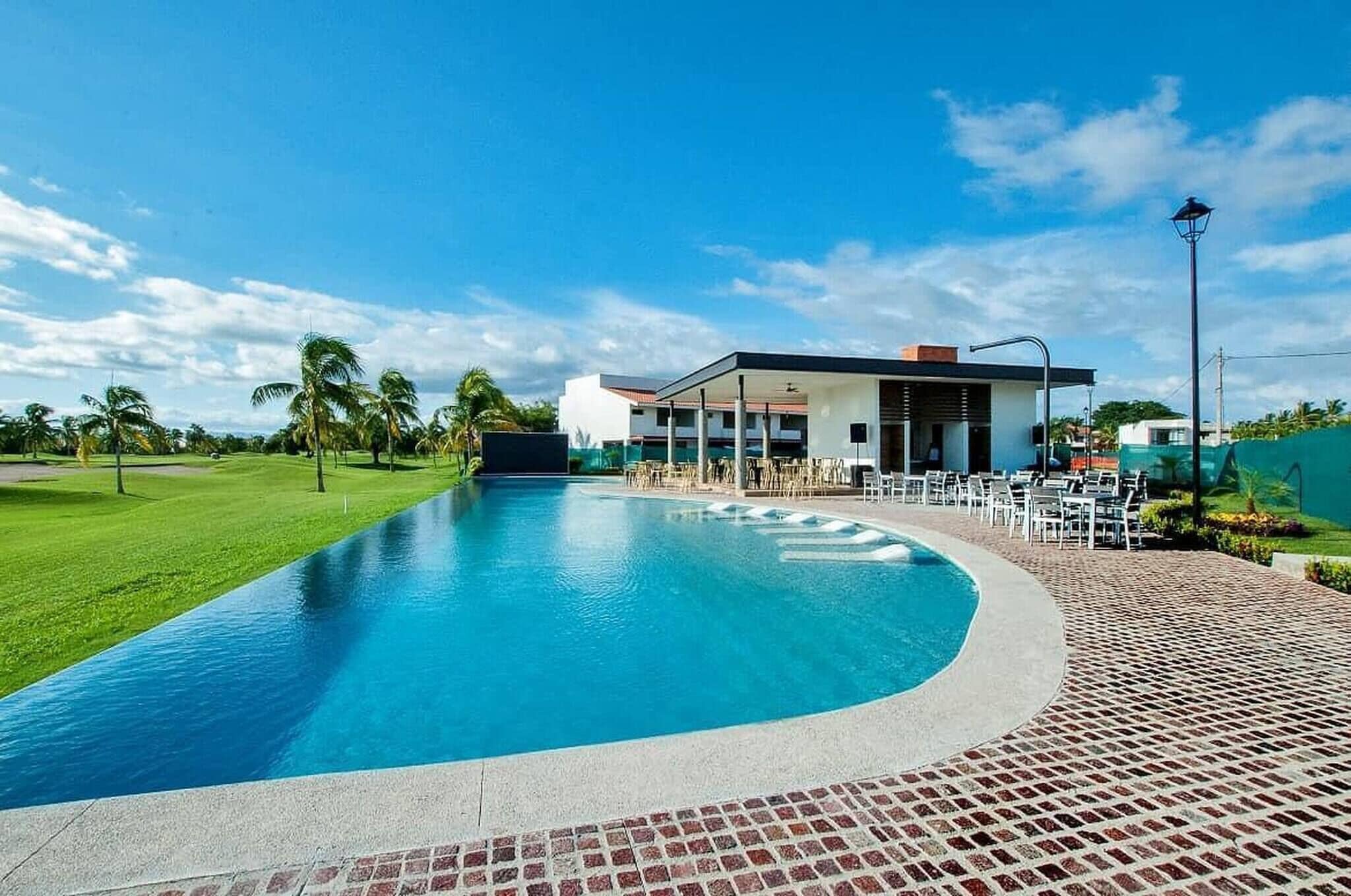 Una piscina con zona de patio, un edificio de techo plano y un cielo azul despejado.