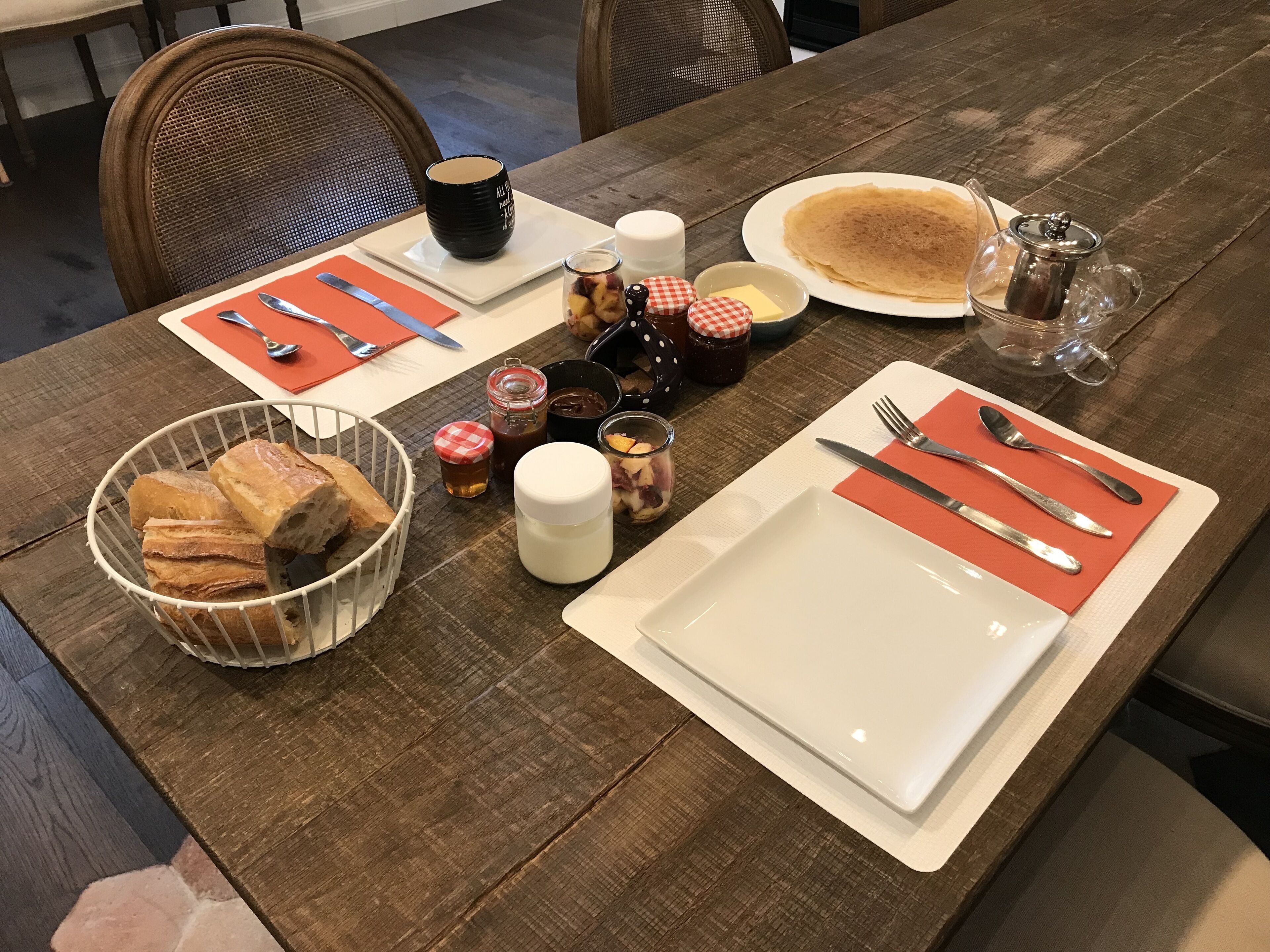 Mesa posta com pratos, talheres, uma cesta de pão, acompanhamentos e uma cafeteira.