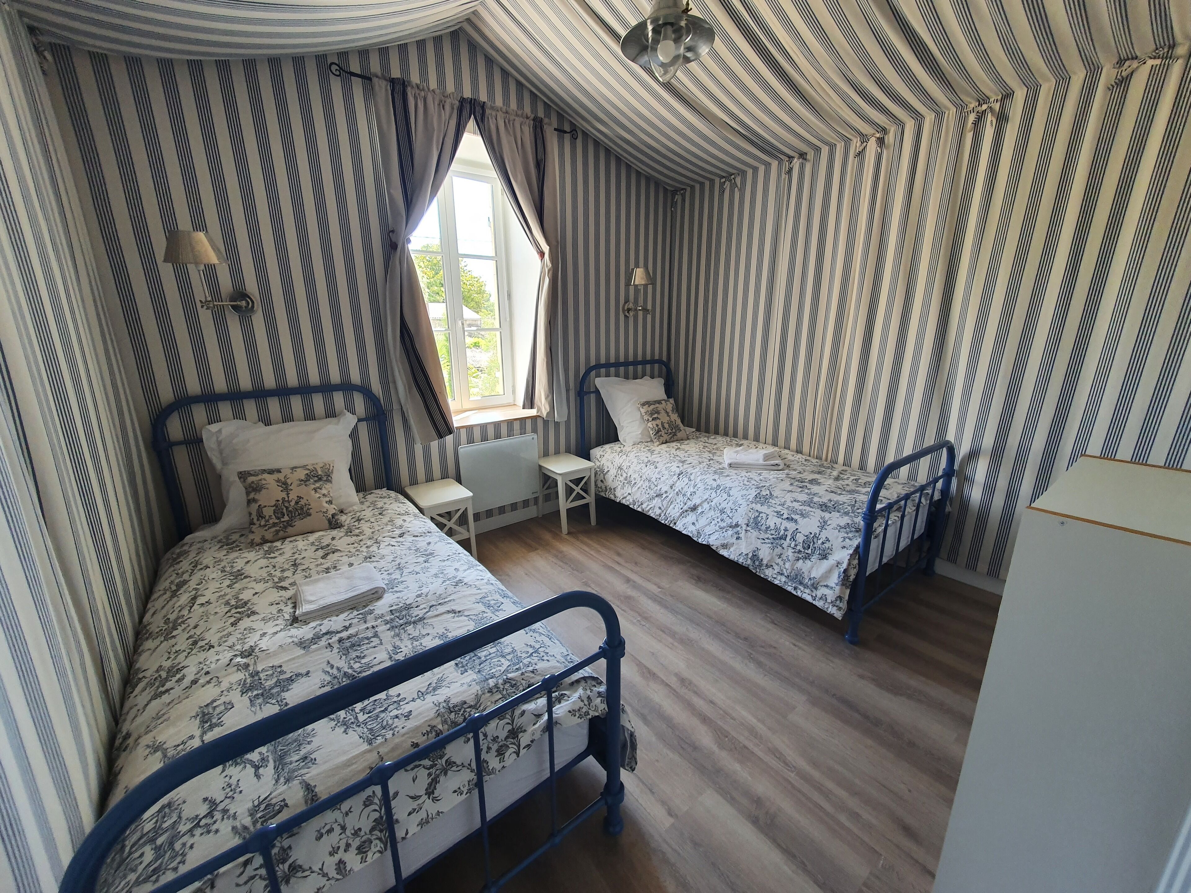 A bedroom with two beds, striped wallpaper, a window, and a ceiling fan.