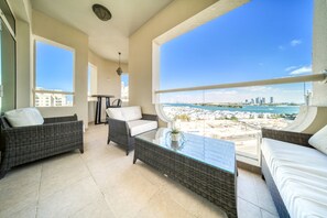 A balcony with wicker furniture, a glass table, and a view of a beach and city skyline.