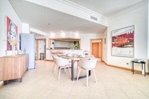 A spacious dining area with a long table, white chairs, and a kitchen in the background.