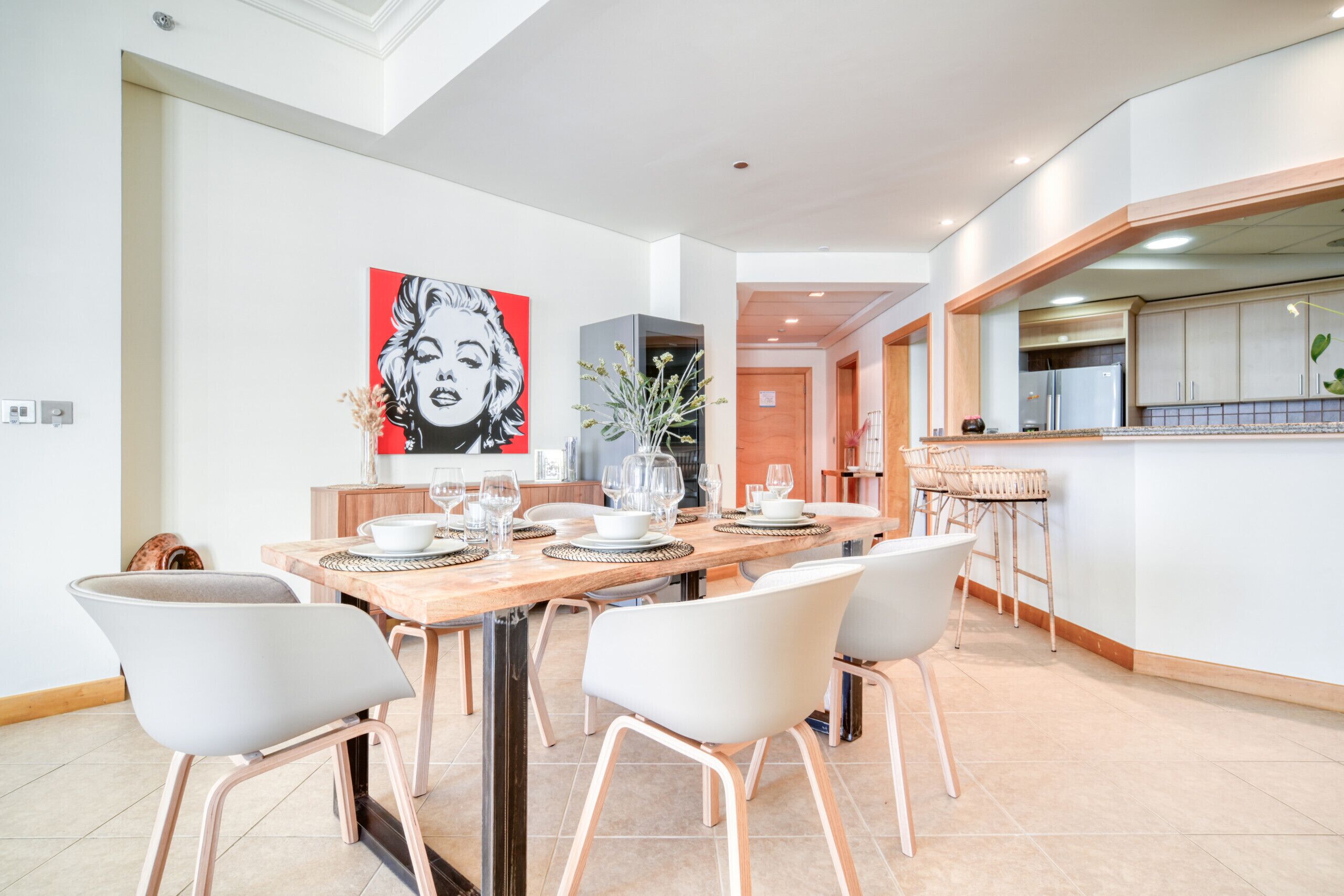 Un comedor moderno con una mesa de madera dispuesta para cuatro, sillas blancas y un cuadro de arte pop grande en la pared.