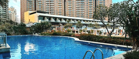 A swimming pool with a modern design, surrounded by lush greenery and a multi-story hotel building in the background.