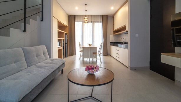 A modern living room with a sofa, a dining table with chairs, a kitchen area, and a staircase. - Anahata Home (Tabanan)