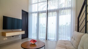 A modern living room with a flat-screen TV, a wooden coffee table with a flower vase, a beige sofa, and large windows with sheer curtains. - Anahata Home (Tabanan)