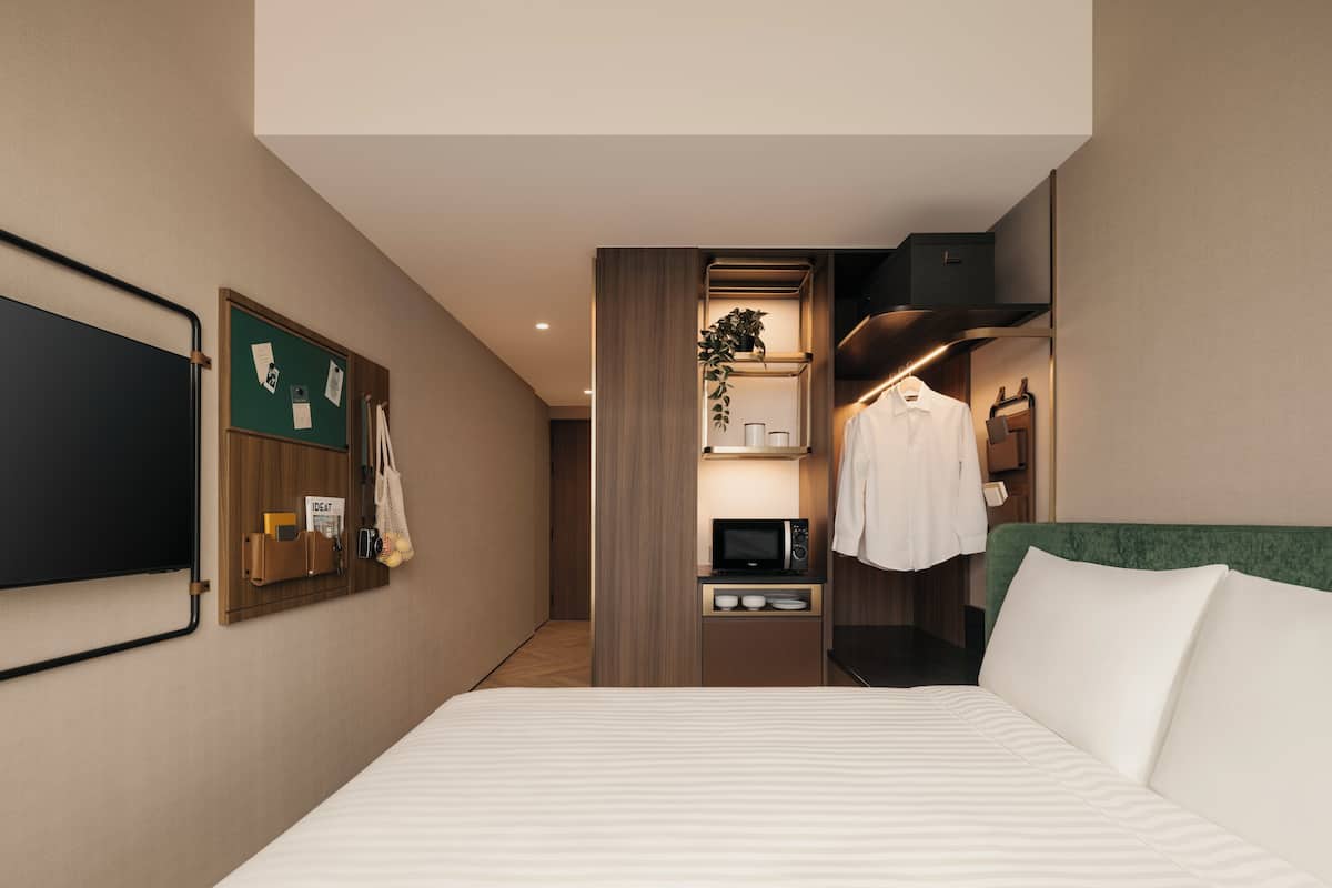 A hotel room with a bed, a desk with a microwave, a hanging plant, and a coat rack.