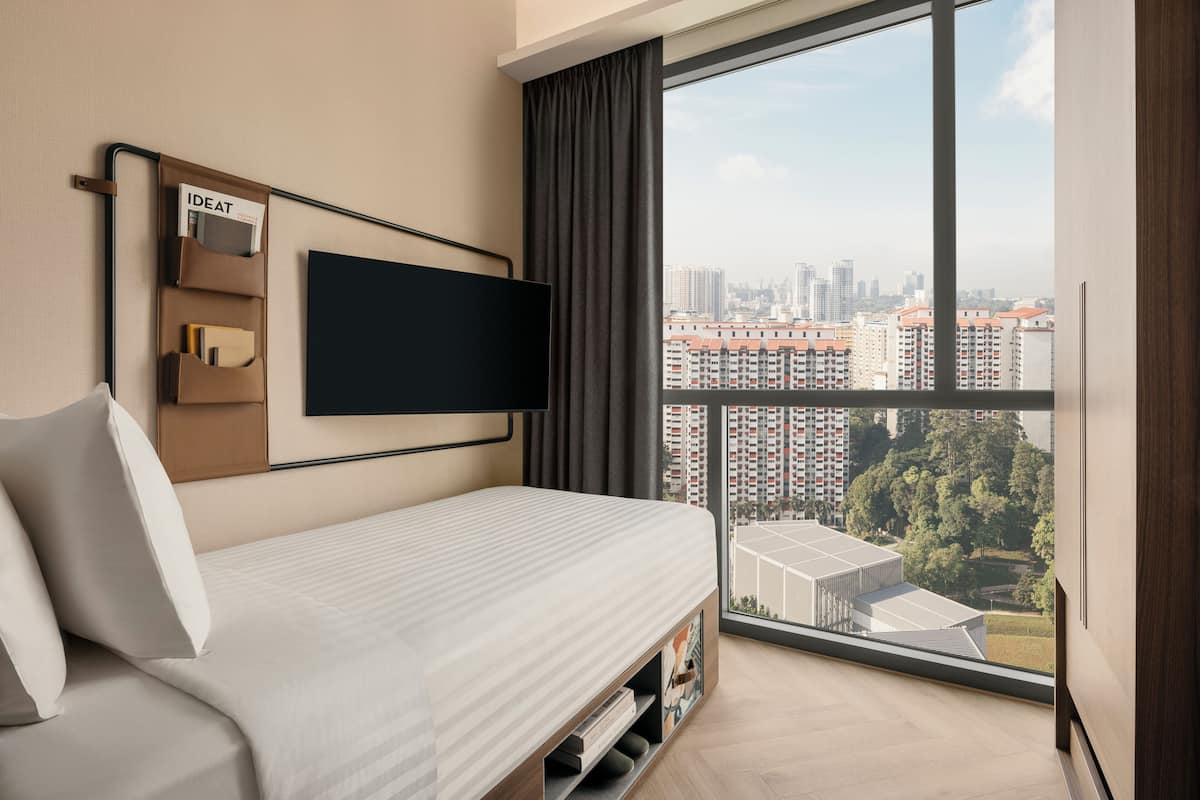 A modern hotel room with a large window, a flat-screen TV mounted on the wall, a bed with a white mattress, and a built-in bookshelf.