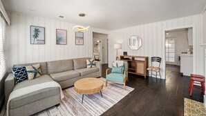 A living room with a sofa, armchair, and round coffee table. There is a desk with a chair and a mirror on the wall. - Silver Surf Bungalow w/ Hot Tub - Steps from Beach (Oxnard)