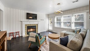 A living room with a fireplace, a television mounted on the wall, and a large window with blinds. - Silver Surf Bungalow w/ Hot Tub - Steps from Beach (Oxnard)