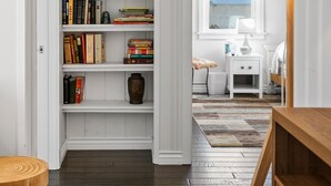 A room with a wooden floor, a white bookshelf, a small white nightstand, and a window with blinds. - Silver Surf Bungalow w/ Hot Tub - Steps from Beach (Oxnard)