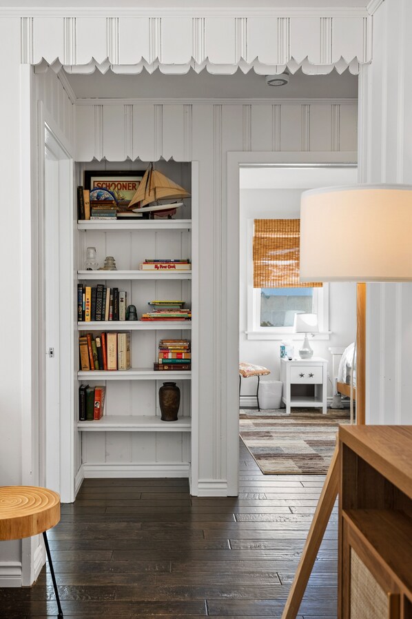 A room with a wooden floor, a white bookshelf, a small white nightstand, and a window with blinds.