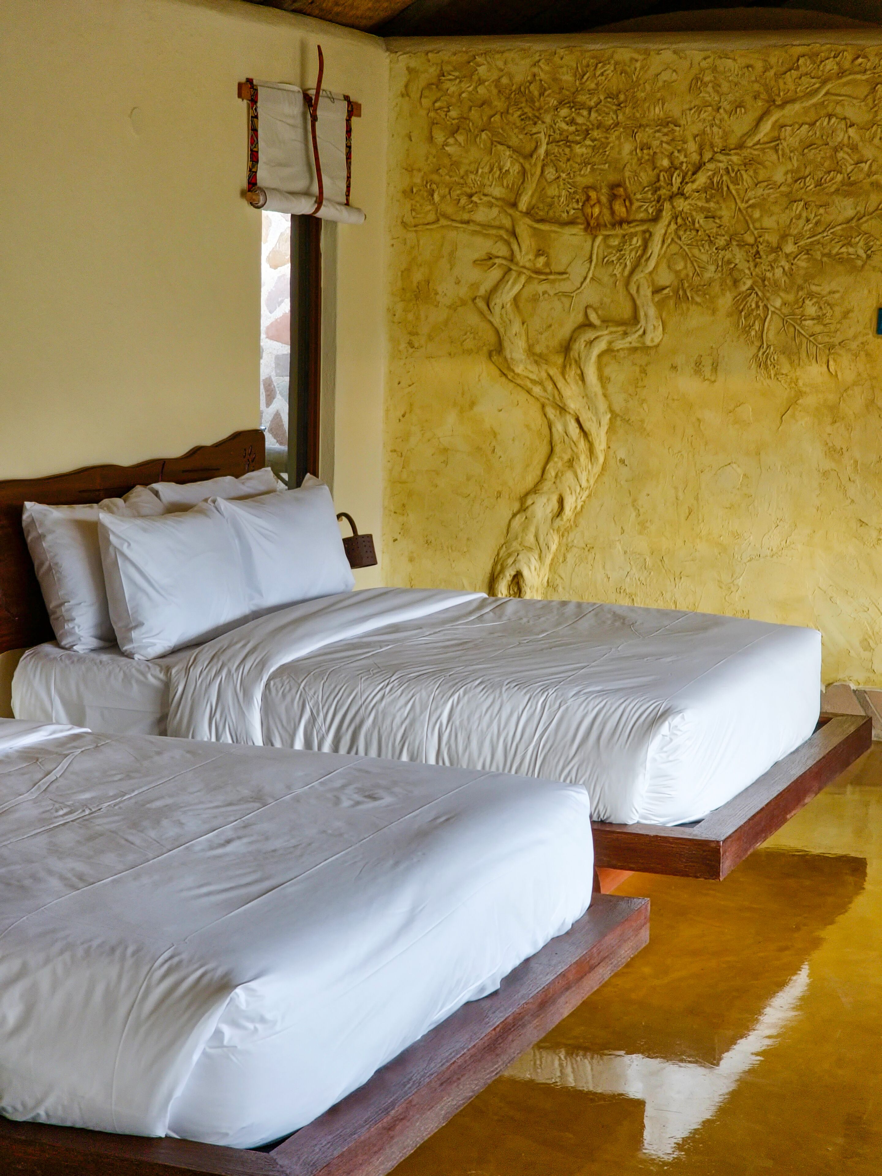 Un lit double avec du linge de lit blanc, dans une pièce dont un mur est peint en jaune avec une texture particulière et dont le sol est en bois.