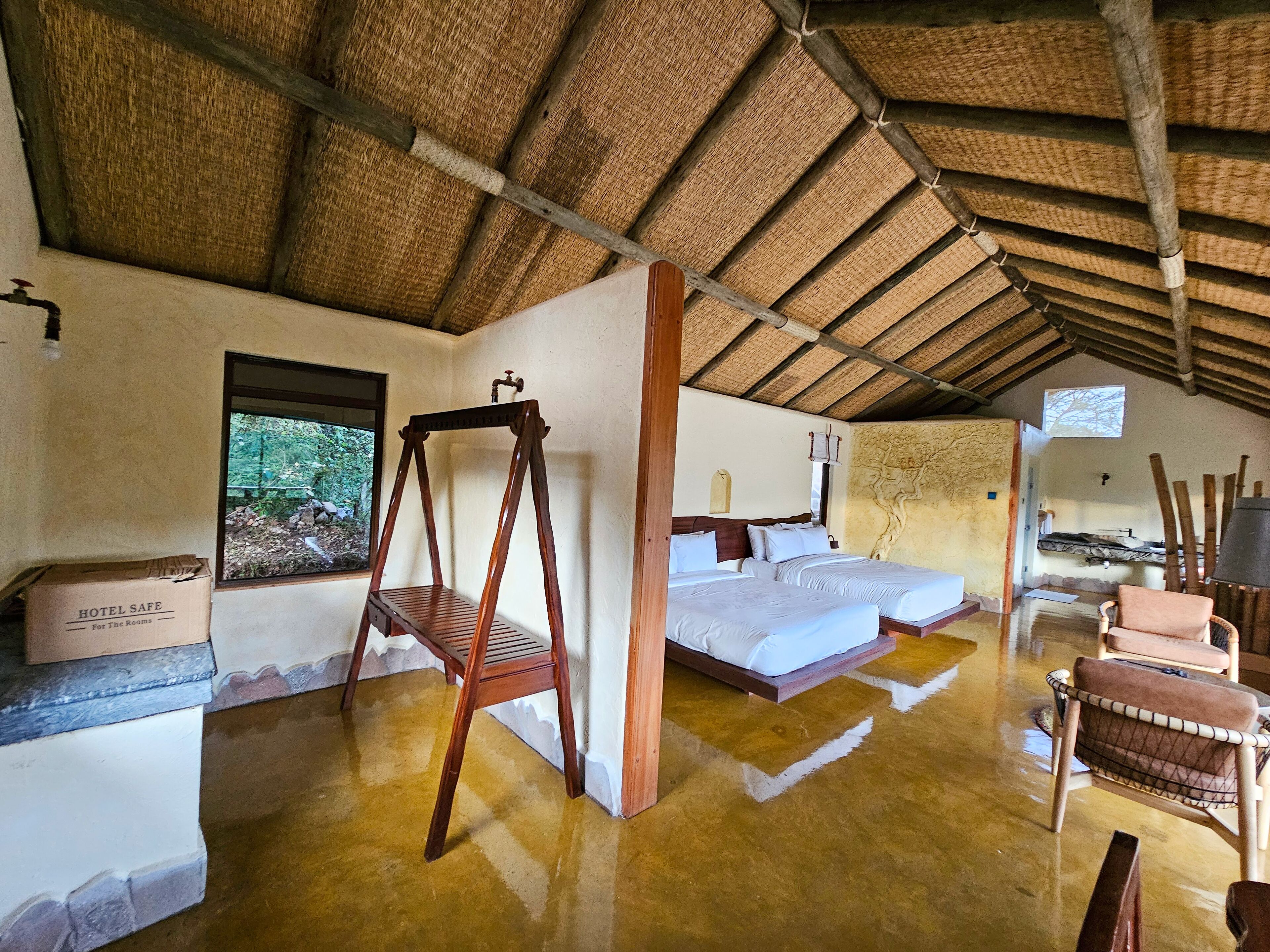 Una habitación rústica con techo de paja, dos camas, una hamaca de madera y vistas al exterior.