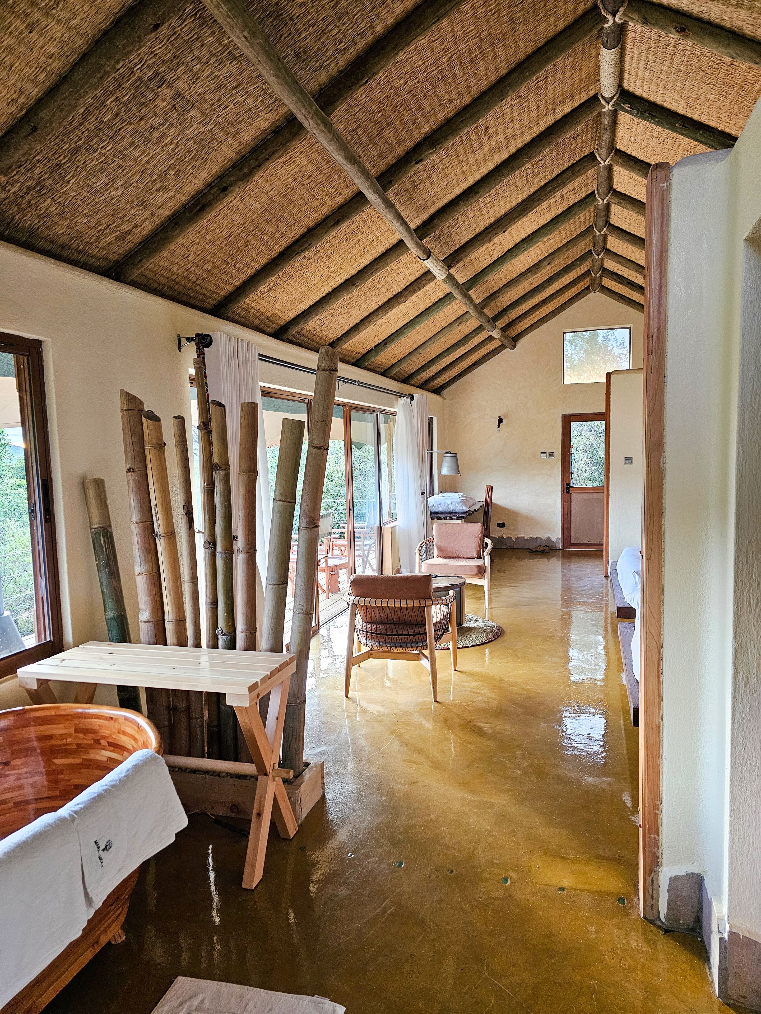 Une pièce spacieuse avec un plafond haut, des poutres en bois et un plancher ciré. On y trouve des poteaux en bambou, une table en bois et un fauteuil rembourré.