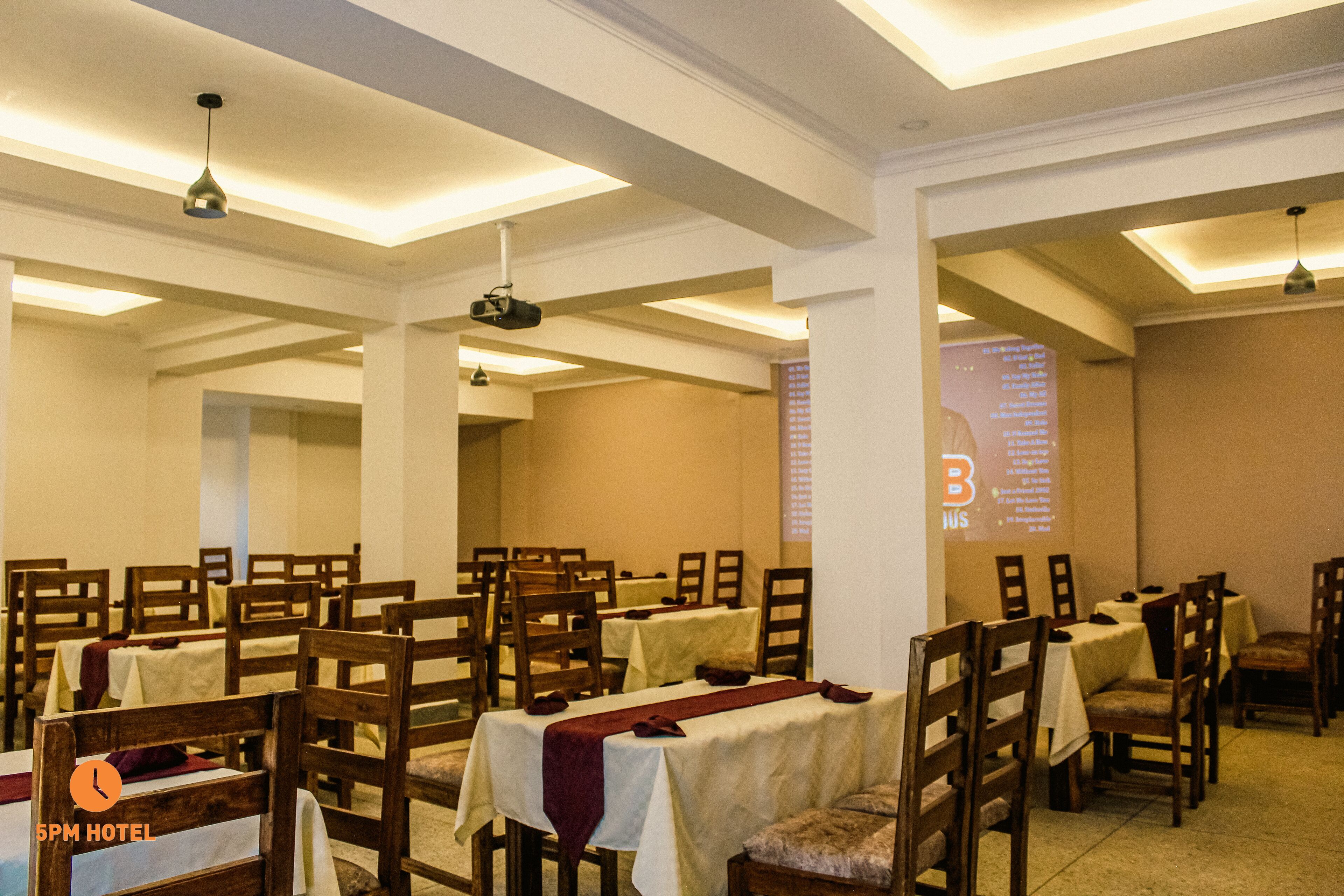 A hotel dining area with tables set for guests, wooden chairs, and a projector screen displaying information.
