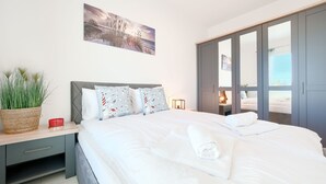 A neatly made bed with white linens, a bedside table with a red mug, and a wall-mounted painting of a beach scene. - Apartamenty Sun & Snow Pelikan (Rewal)