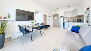 A modern living room with a grey sofa, a dining area with a table and chairs, a kitchen with a fridge and microwave, and a large window. - Apartamenty Sun & Snow Pelikan (Rewal)