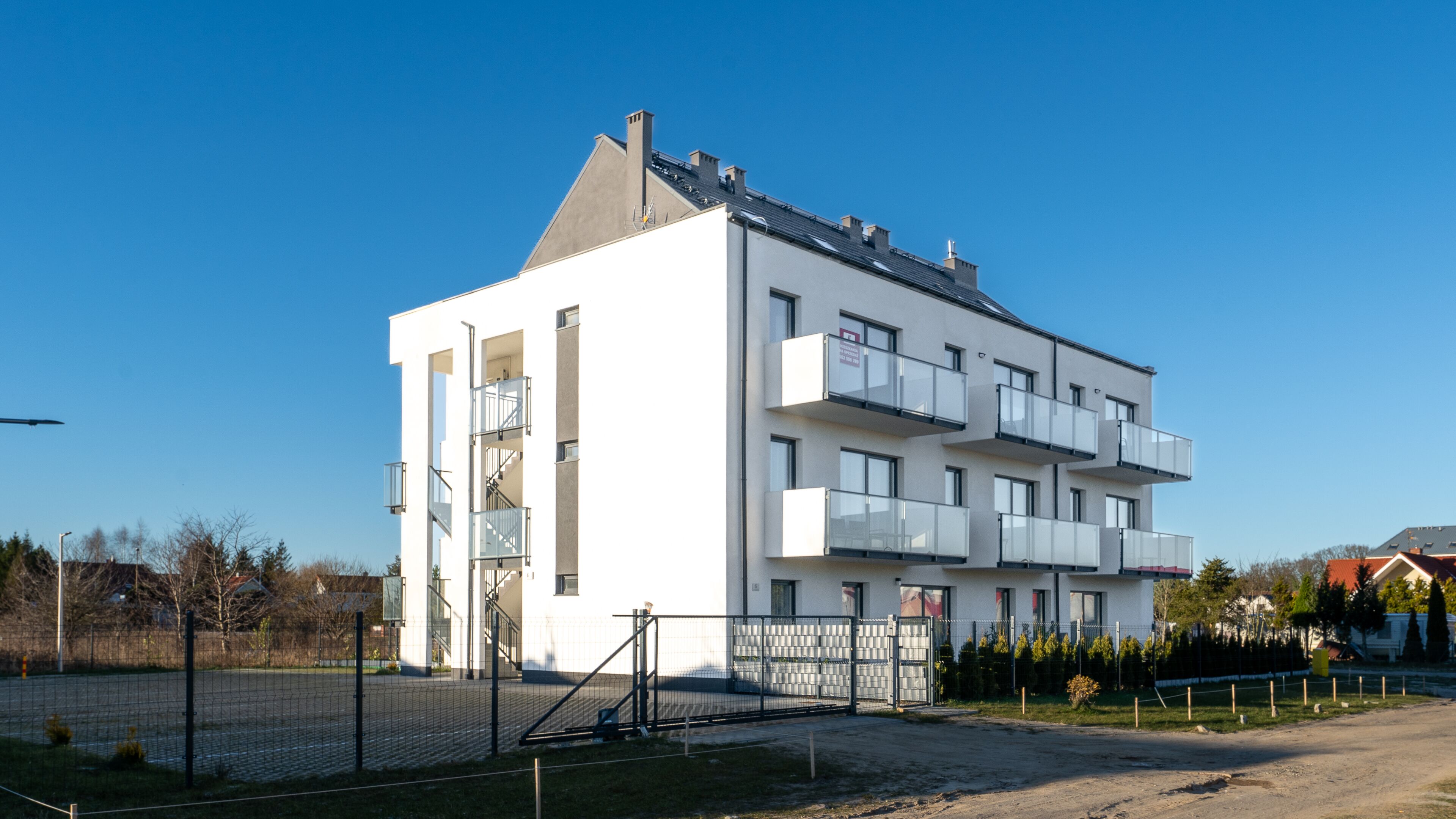 A modern multi-story residential building with balconies, a gated entrance, and a clear sky.
