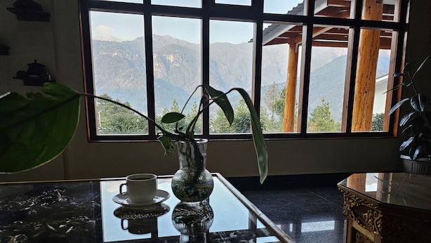 Une pièce dotée d’une grande fenêtre donnant sur les montagnes, d’une table avec une tasse et d’un vase contenant une plante, ainsi que d’un meuble décoratif en bois.