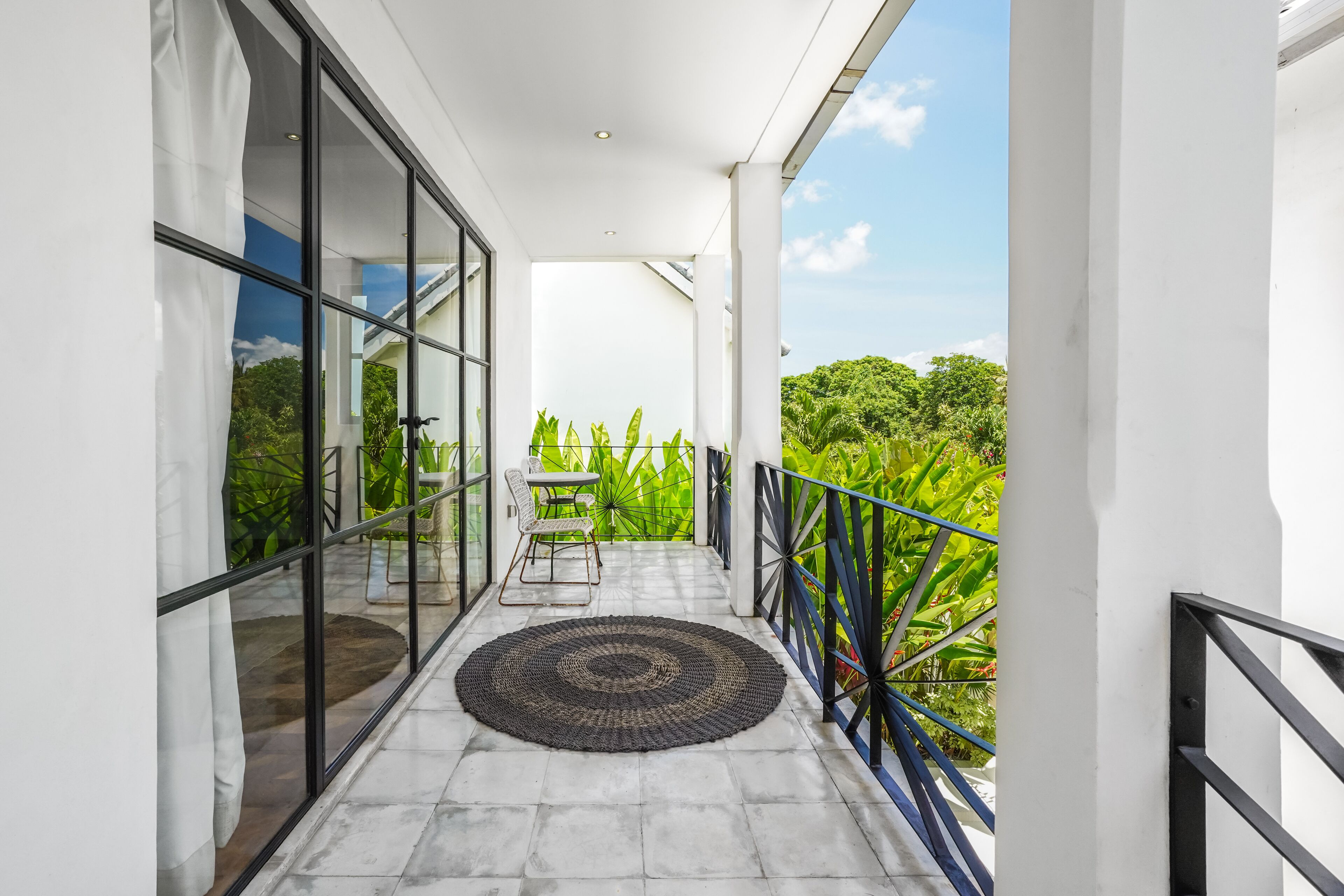Uma varanda com vista para vegetação exuberante, um tapete redondo de fibra natural e uma mesa com cadeiras.