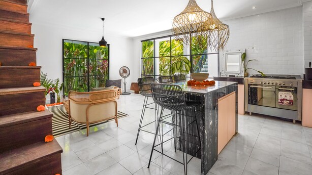 A modern kitchen with a central island, bar stools, a large window with a view of greenery, and a wooden staircase. - Mare Villa by Betterplace (Canggu)