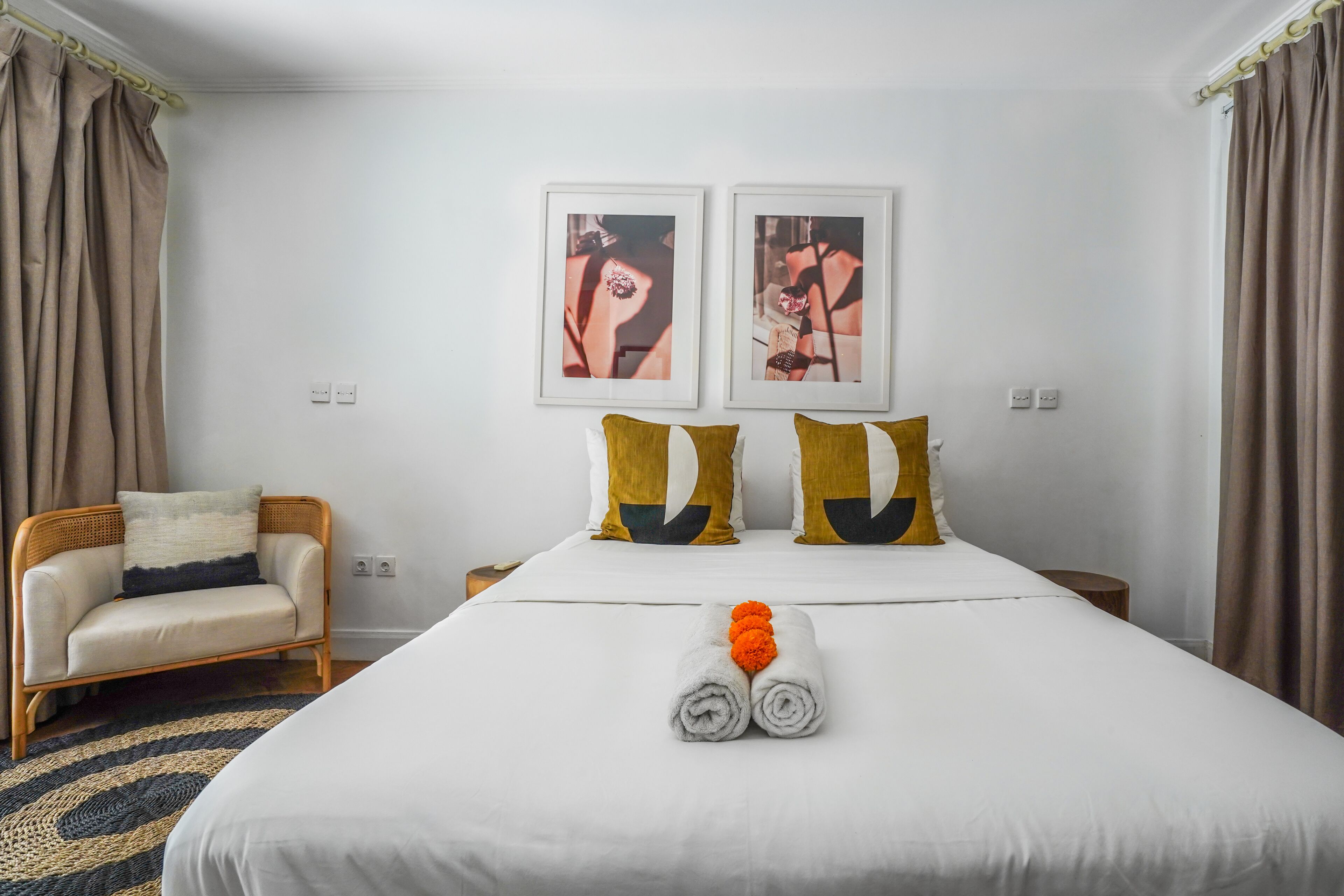 A neatly made bed with white linens, two pillows with yellow and black patterns, and a pair of rolled towels with orange flowers.